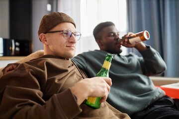 Portrait of two young men drinking beer while sitting on couch at home and watching TV together