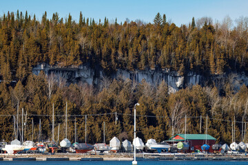 Drydocked boats in the harbor at a marina covered and winterized for the winter. Below white rocky cliffs, trees, and a blue sky.