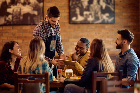 Happy waiter showing the menu on touchpad to group of guests in bar.
