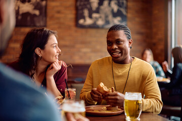 Happy black man eating burger while gathering with friends in bar.