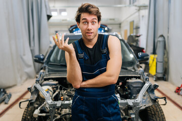 Portrait of troubled handsome mechanic male in uniform looking at camera not knowing what disrepair during maintenance car in auto repair workshop. Doubtful repairman trying to solve problem in garage