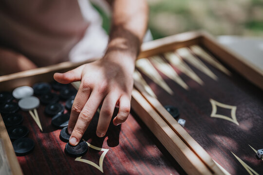Close-up of a person's hand playing backgammon outdoors, capturing the strategic move in a sunlit setting on a wooden board.