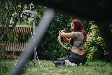 A girl with red hair carefully handles a large snake in a lush green garden. The image captures the...