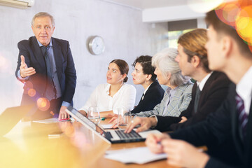 Angry elderly male boss scolding office workers of different ages using notebooks at table in office