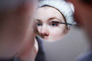 Girl applying make up (paint her eyelashes) in front of a mirror. Focus on her reflection