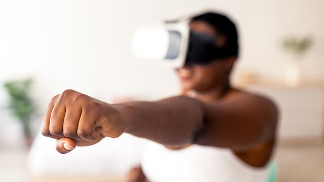 Virtual training concept. Overweight black lady in VR glasses using fighting simulator, making punch at home, focus on clenched fist. Plus size Afro woman boxing with augmented reality app - Powered by Adobe