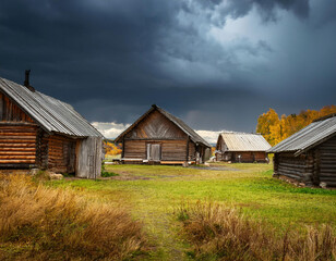 Traditional wooden log cabins stand against a dramatic stormy sky with autumn trees in the background.