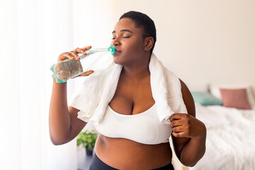 Healthy living, weight loss. Positive plus size black lady drinking fresh clear water from bottle after sports workout indoors. Curvy millennial woman staying hydrated during home fitness exercises