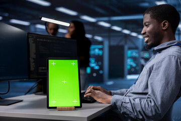 Smiling man working on green screen tablet in modern data center. Happy IT specialist doing IT infrastructure management on server racks in high tech environment using chroma key device