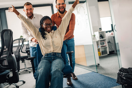 Business colleagues pushing businesswoman on office chair, having fun at work