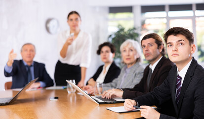 Portrait of young office worker sitting at table with colleagues, listening with interest and watching presentation during business meeting