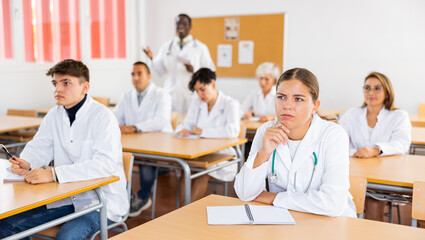 Interested young female student in white coat listening to lecture and taking notes in classroom during professional medical training