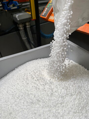 pouring of a bag of white plastic pellets on a gray plastic drawer, production of plastic parts, plastic injection, spilling of plastic pelets, with a plastic injector out of focus in the background