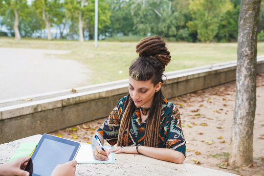 An agender person, distinguishable by their dreadlocks and fringe, engages in writing
