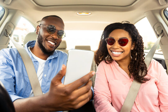Portrait of smiling African American couple wearing sunglasses driving luxury car, male driver holding and using mobile phone, showing road to woman, looking at digital map application