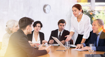 Young European woman stadning in front of office workers sitting using laptops and making notes at table in meeting room