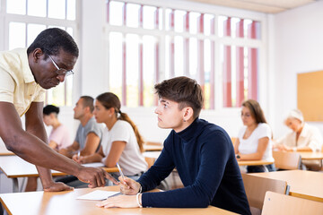 Fototapeta premium Strict African American teacher standing next to focused young guy sitting at desk during lesson, pointing out error in solving task in student notebook..