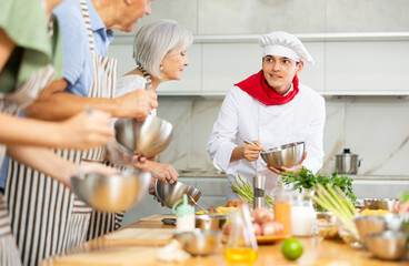 Enthusiastic young chef giving culinary classes to group of interested men and women of different ages, teaching to cook deliciously..