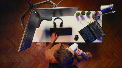 Top down view of engineer doing multitasking to finish developing PC code, drinking coffee, listening music. IT admin wearing headphones, solving database errors on computer and laptop, aerial shot