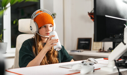 Medium shot of young student girl in headset sitting at computer in cyberspace, playing intense video game, finishing level, leaning back on chair and sipping energy drink from can. 