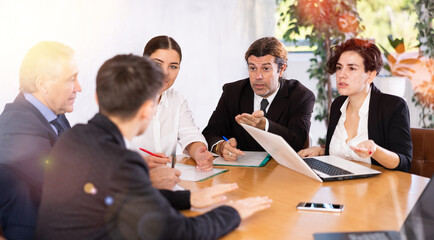 Office coworkers of different ages discussing topic of day while sitting at table with laptop and notes