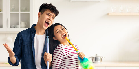 Joyful asian loving couple singing songs while cleaning kitchen, using fluffy dust brush as...