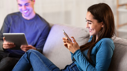 Modern Technologies Concept. Beautiful spouses sitting on couch in their apartment, using digital tablet and smart phone