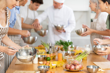 Pleased young woman member of cooking club trying to prepare sauce standing around kitchen table...