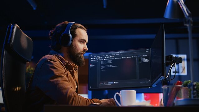 Cheerful programmer listening music while doing web development during remote job shift in neon lit home office, finishing coding project. IT expert enjoying audio podcast, working on PC, camera B