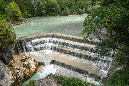 A scenic view of a Lechfall waterfall in Fussen, Bavaria, cascading over a stepped dam into a river, surrounded by lush green trees and a serene landscape.