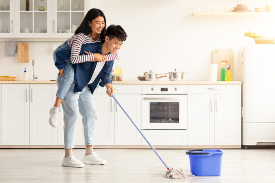 Cheerful asian guy piggyback his pretty girlfriend while mopping floor at cozy white kitchen, panorama with copy space. Loving korean couple having fun while house-keeping their new apartment - Powered by Adobe