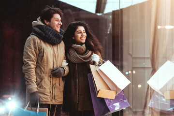 New Year Sales. Couple On Winter Shopping Holding Shopper Bags Looking At Clothing Store Window In Night City.