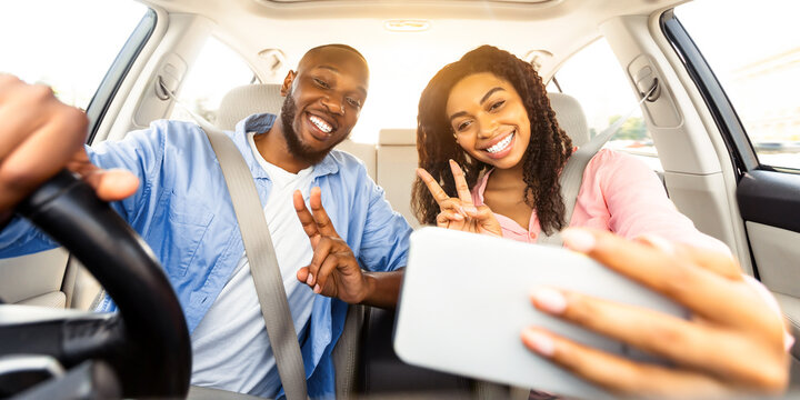 Beautiful young African American couple taking selfie inside car, using mobile phone, showing v victory peace sign gesture. Happy black man and woman going on vacation, taking self photo in auto
