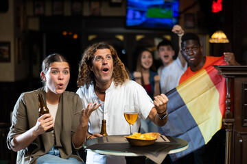 Emotional multiracial sport supporters waving flag of Belgium and resting with beer and chips in the sport bar