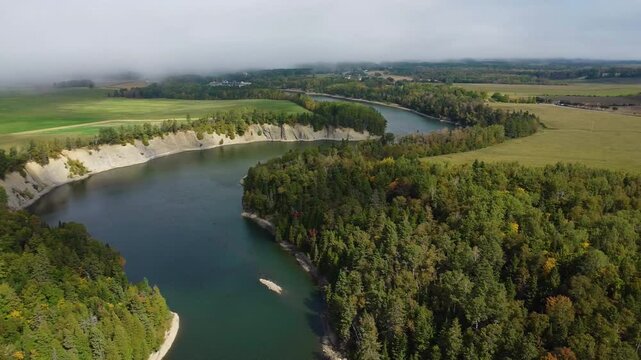 Rotating panoramic aerial view of a winding river emerging from low clouds and its rocky cliffs surrounded by agricultural fields and a rural village. Mitis River, Price, Quebec.