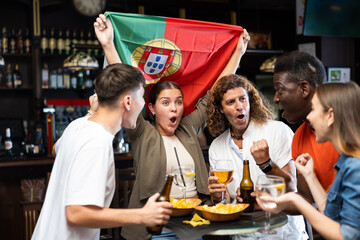 Group of cheerful emotional people of different ages and nationalities cheering for favorite sports team in pub, waving national flag of Portugal while watching football match on TV..