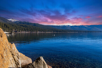 a gorgeous winter landscape at Lake Tahoe with blue lake water, lush green trees and plants and snow capped mountains at sunset in Crystal Bay Nevada USA