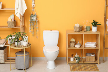 Interior of stylish restroom with white toilet bowl, coffee table and shelving unit near orange wall