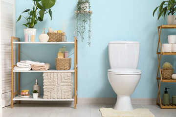 Interior of stylish restroom with ceramic toilet bowl, houseplant, supplies and shelving unit near...