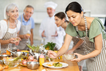 Attentive middle-aged woman attendee of cooking course decorating meal made of salmon fish together...