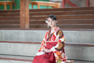 Japanese woman in her 20s wearing red hakama, staple for Japanese university graduates, sits in audience seats in large room.