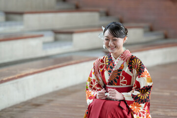 A teenage man wearing casual clothes uses a selfie camera to photograph a Japanese woman in her 20s wearing a red hakama, a staple for Japanese university graduates.