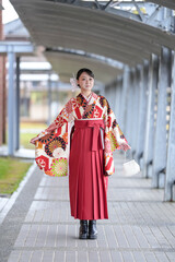 A Japanese woman in her 20s, wearing a red hakama, a staple for Japanese university graduates, poses cutely in front of a cultural building.