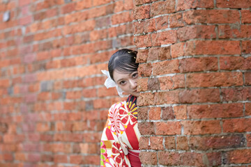 A Japanese woman in her 20s wearing a red kimono (Hakama), a staple of Japanese university graduates, stands in front of a cultural brick building, peeking out from the pillars.