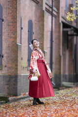 Obraz premium A Japanese woman in her 20s, wearing a red kimono (hakama), a staple of Japanese university graduates, dances on a path filled with autumn leaves in front of a traditional Japanese brick building.
