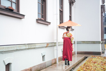 A Japanese woman in her 20s wears a red kimono (hakama), a staple of Japanese university graduates, and stands holding a red Japanese umbrella in front of a traditional Japanese whitewashed building.