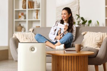 Modern air purifier against woman with magazine sitting on sofa at home, closeup