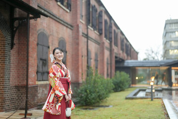 Fototapeta premium A Japanese woman in her 20s stands in the rain in front of a traditional brick building wearing a red kimono (hakama), a staple of Japanese university graduates.