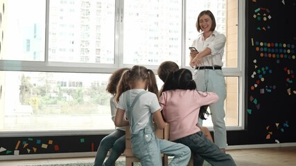 Diverse children playing musical chairs games while young smart beautiful teacher turn off music. Multicultural students running and sitting on chair in creative activity. Music lesson. Erudition.