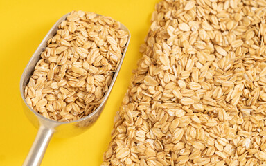 Metal scoop filled with rolled oats beside a scattered pile on a yellow background. Ideal for healthy eating and natural food concepts.